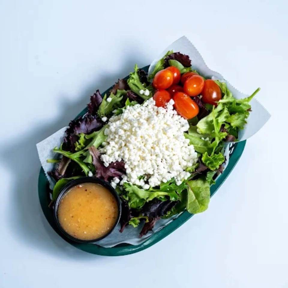 A fresh green salad with cherry tomatoes, crumbled feta cheese, and a side of dressing.